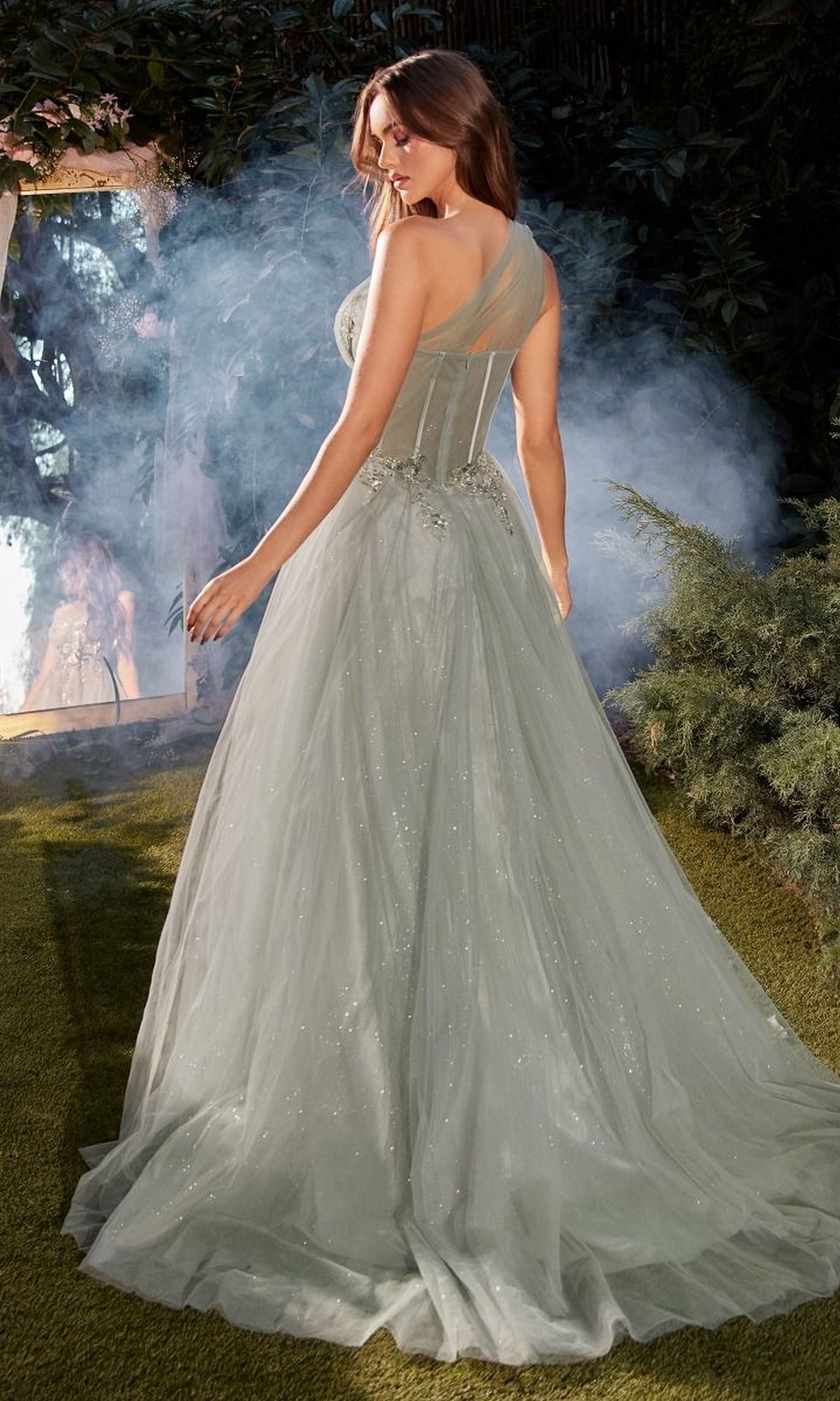 Woman wearing the Sage Green Long Prom Gown (Andrea and Leo A1259) stands outdoors on grass near smoke, facing away, with her reflection visible in a mirror.
